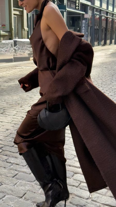 Model in a long brown coat striding over cobblestones with the black bag