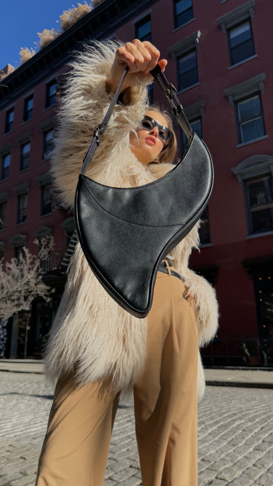 Model raising the Made in Neptune crescent bag in a cream faux-fur coat on a New York street