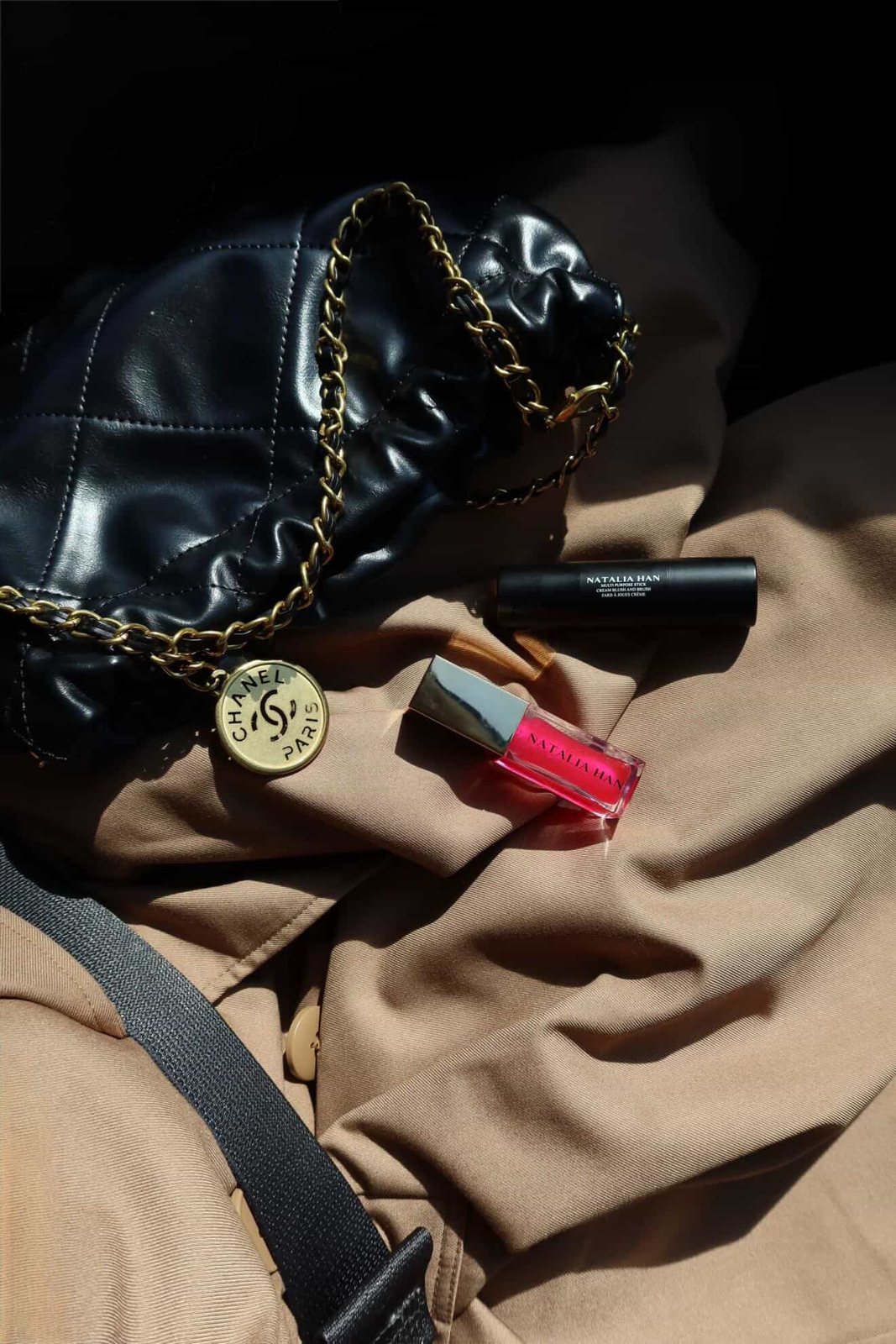 Natalia Han Beauty blush stick and lip oil styled with a quilted black bag on a camel coat in hard sunlight